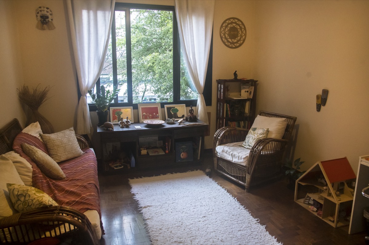 A sala do consultório, com sofá, poltrona de vime, tapete e janela para as árvores